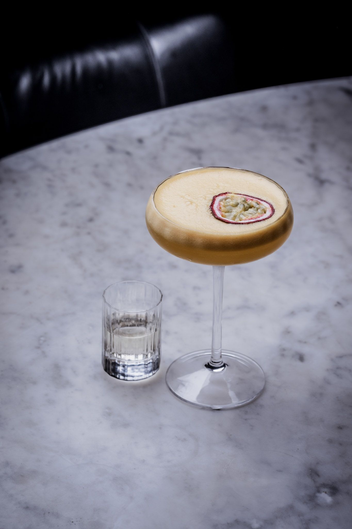 Cocktail in a stemmed glass with a frothy top, passion fruit garnish, and a small shot glass on a marble table.