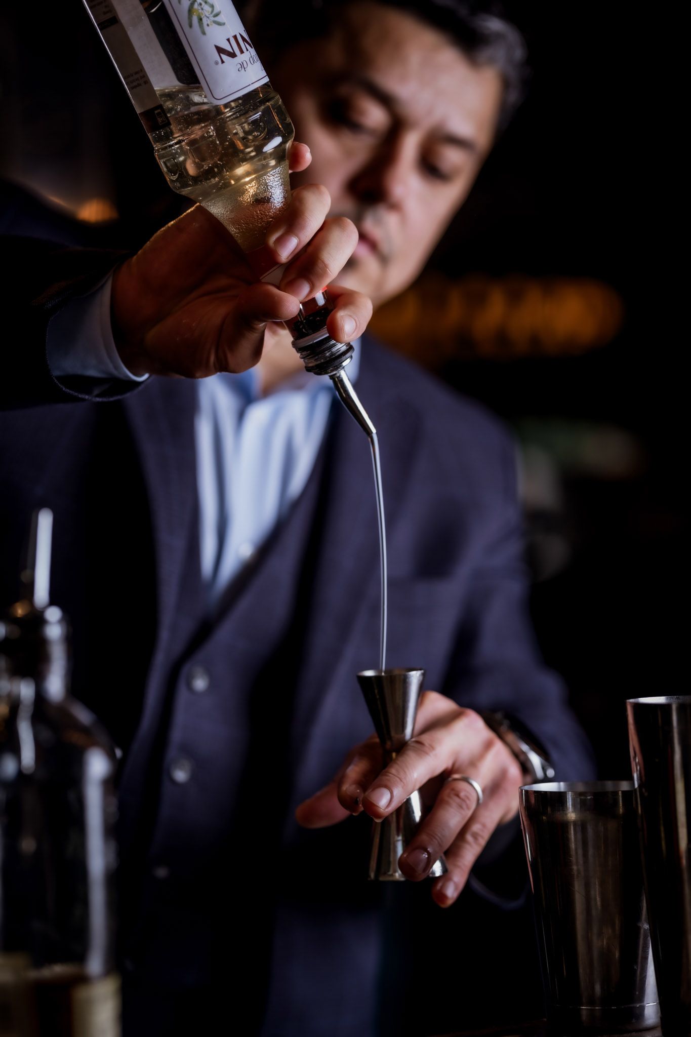Bartender pouring liquid from a bottle into a jigger, preparing a cocktail. Dark setting.