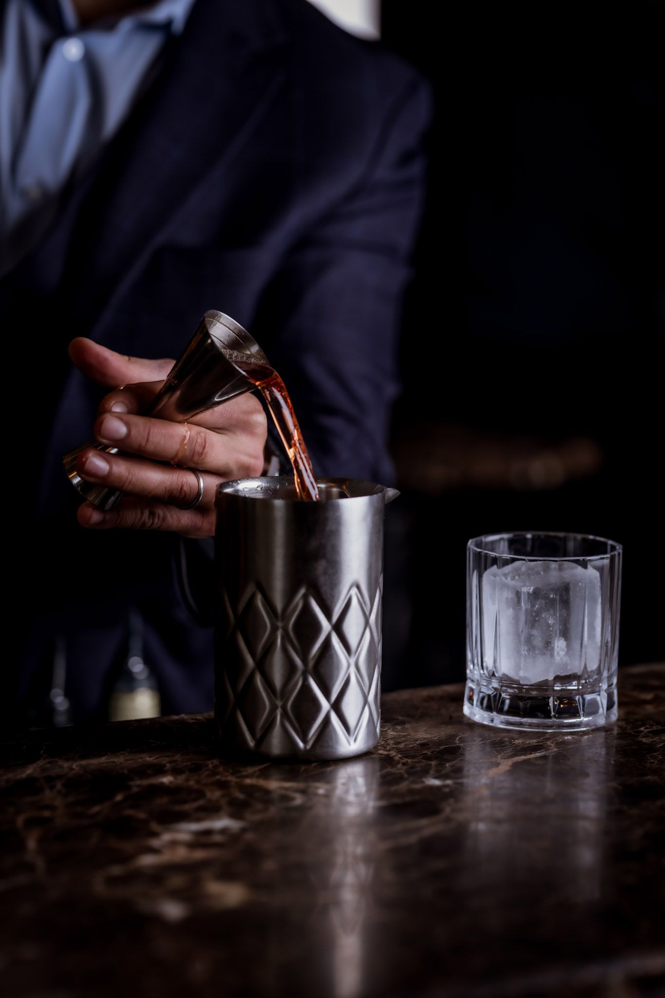 Bartender pouring a drink from a jigger into a shaker, next to a glass with an ice cube.