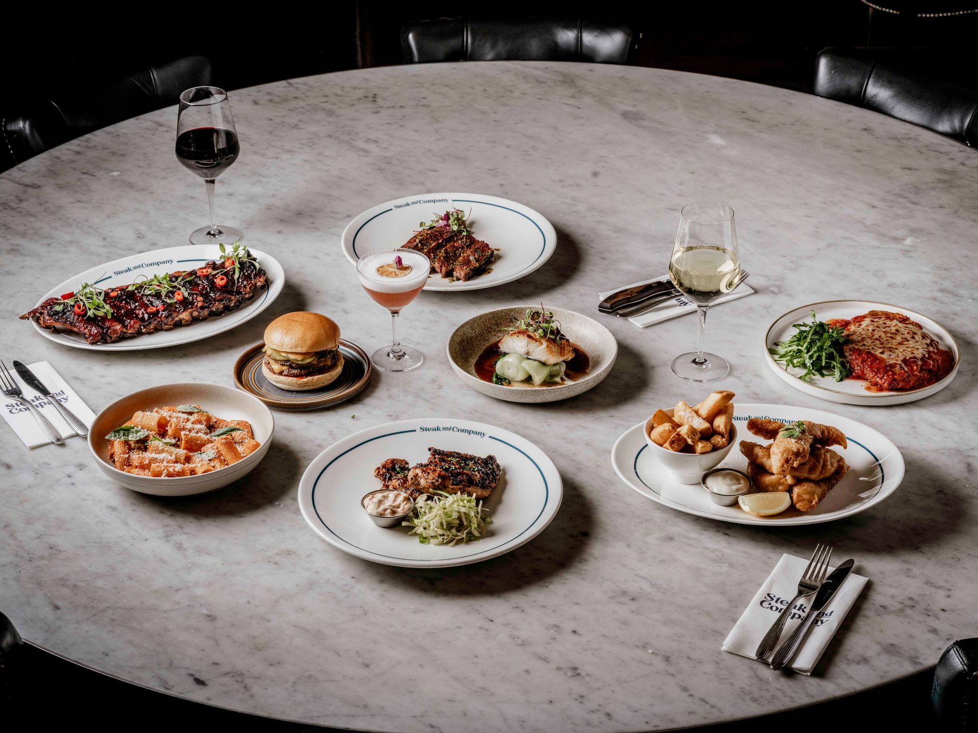 Main dishes of Vegan Steak, BBQ Ribs, Fish and Chips, Miso Black Cod, Chicken Parmigiana, a burger and two glasses of wine on a light marble table.