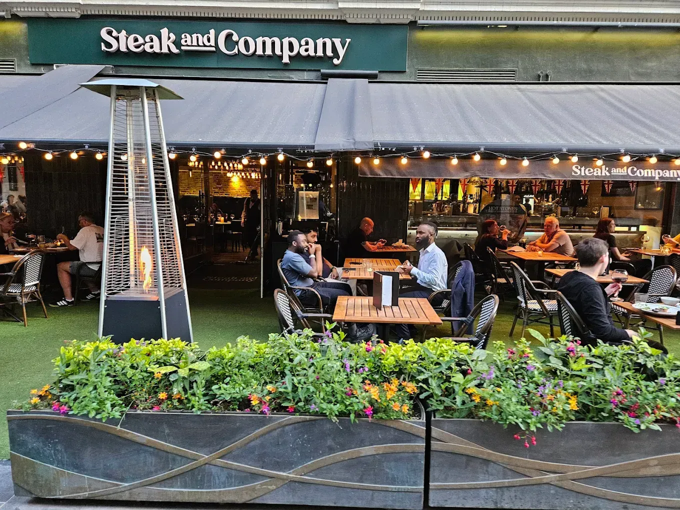 Steak and Company restaurant exterior with outdoor seating. People dine at tables on a patio with a heater.
