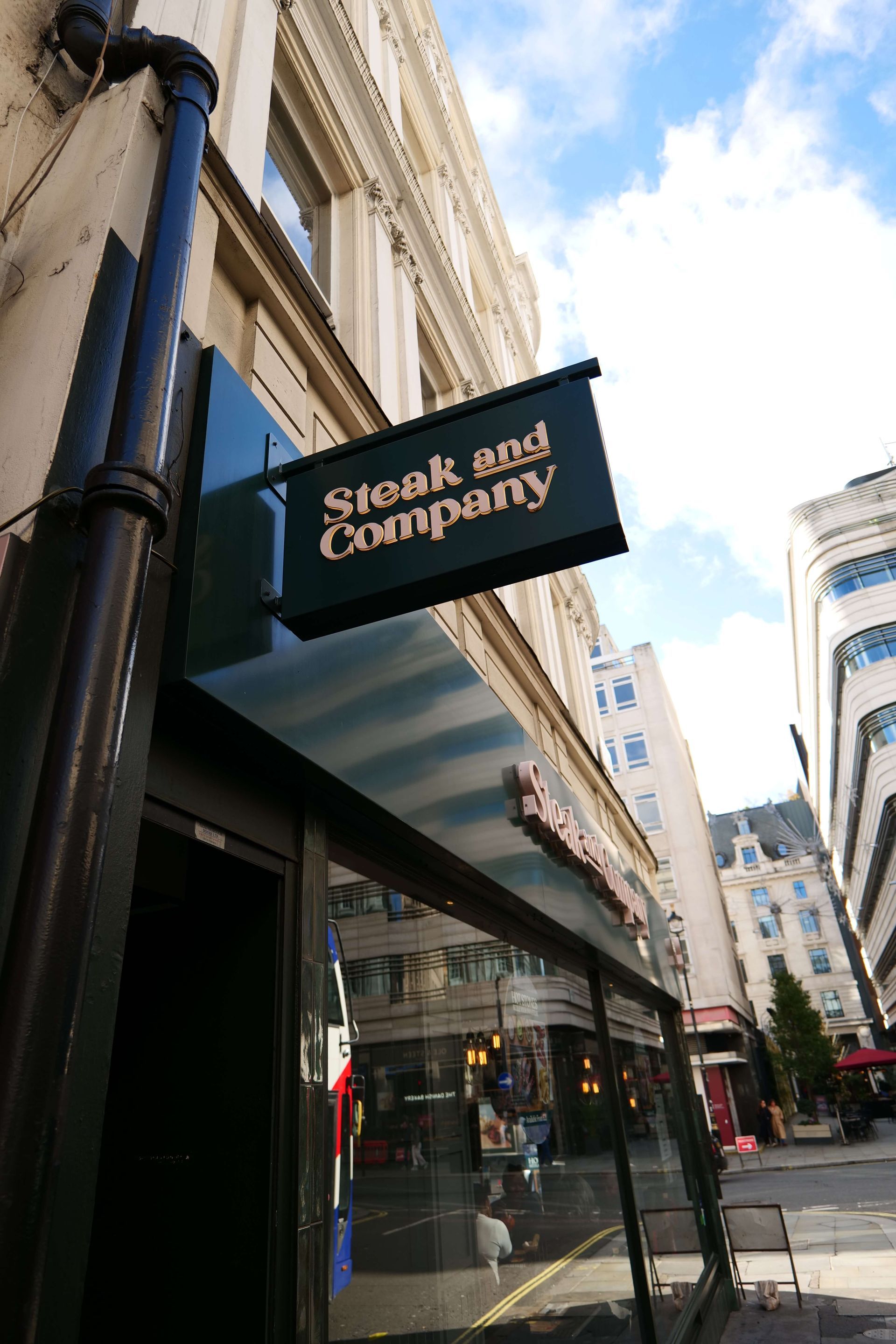 Steak and Company restaurant exterior. Green facade, large windows, outdoor seating, and a busy street scene.