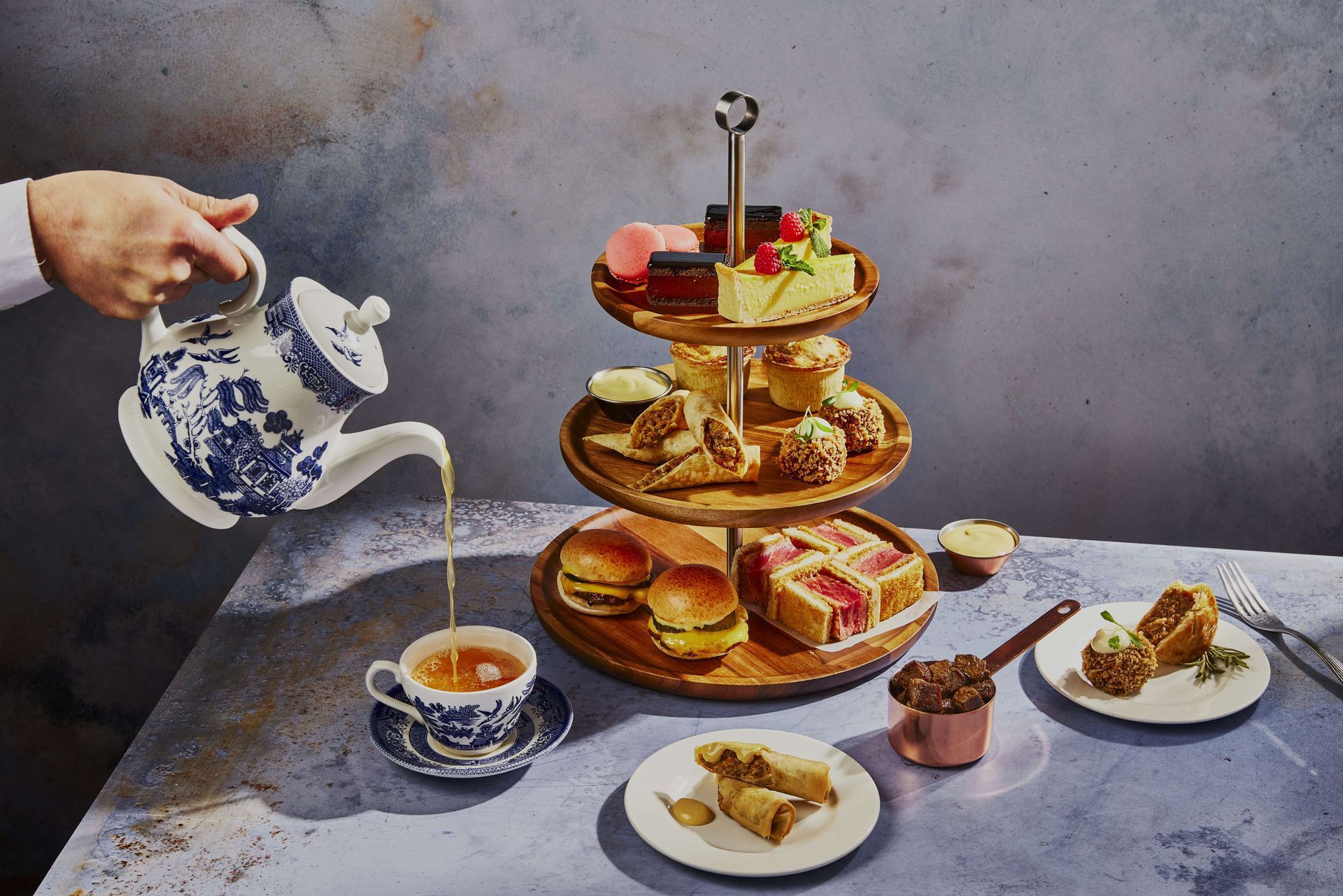 A hand pours tea into a blue and white cup. A tiered wooden server holds desserts like cakes and sandwiches.