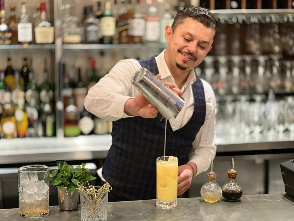 Bartender pouring a drink from a shaker into a tall glass at a bar.