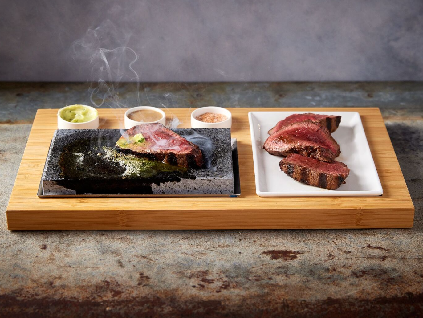 Steak on a hot stone with condiments and cooked slices on a wooden board.