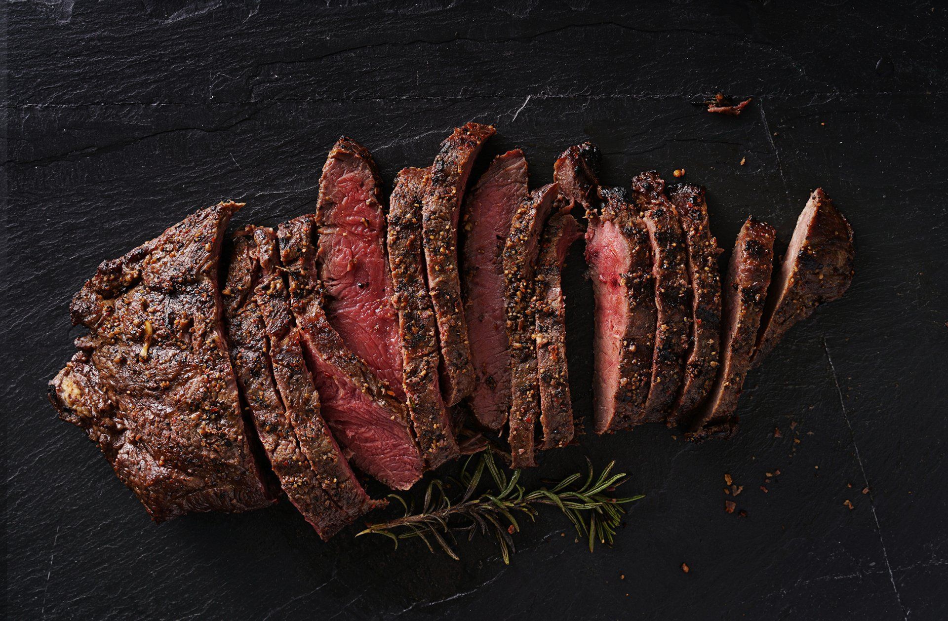 Sliced, cooked steak on a dark surface, with a sprig of rosemary.