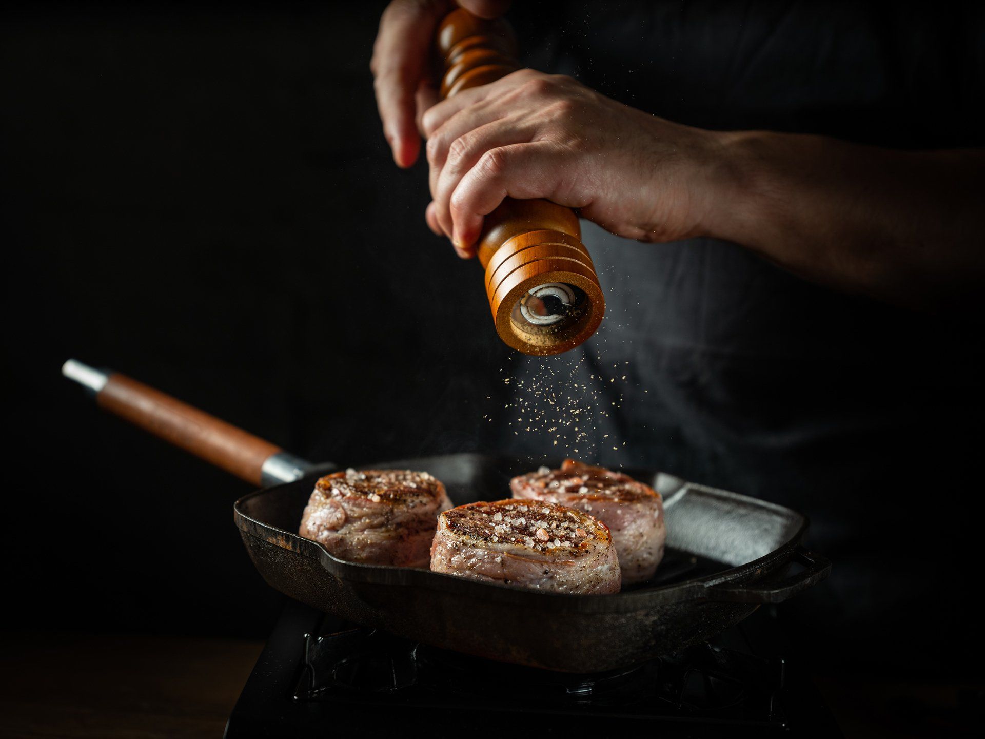 A person seasoning three seared steaks in a cast iron pan with a pepper grinder.