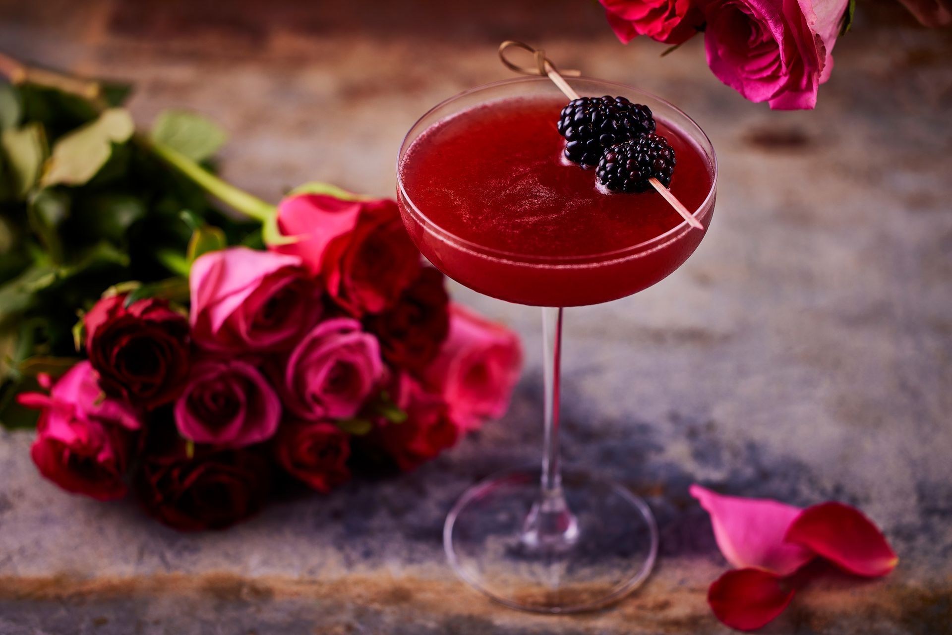 Cocktail in a stemmed glass garnished with blackberries, beside a bouquet of pink roses.
