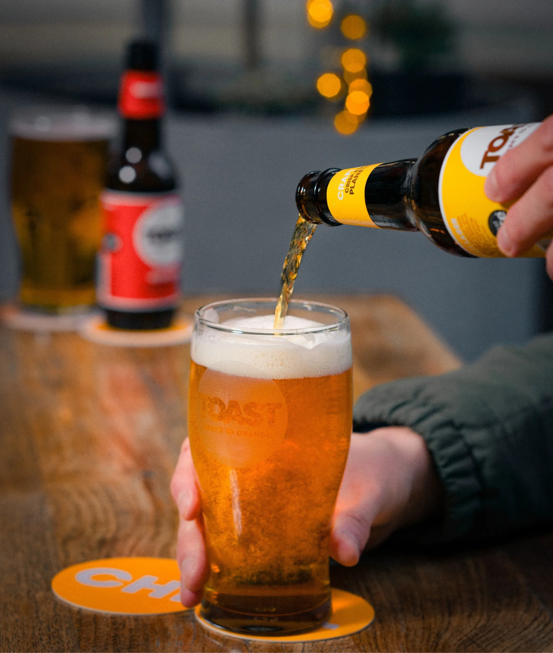 Beer being poured from a bottle into a glass, on a wooden table. Other beer bottles visible.