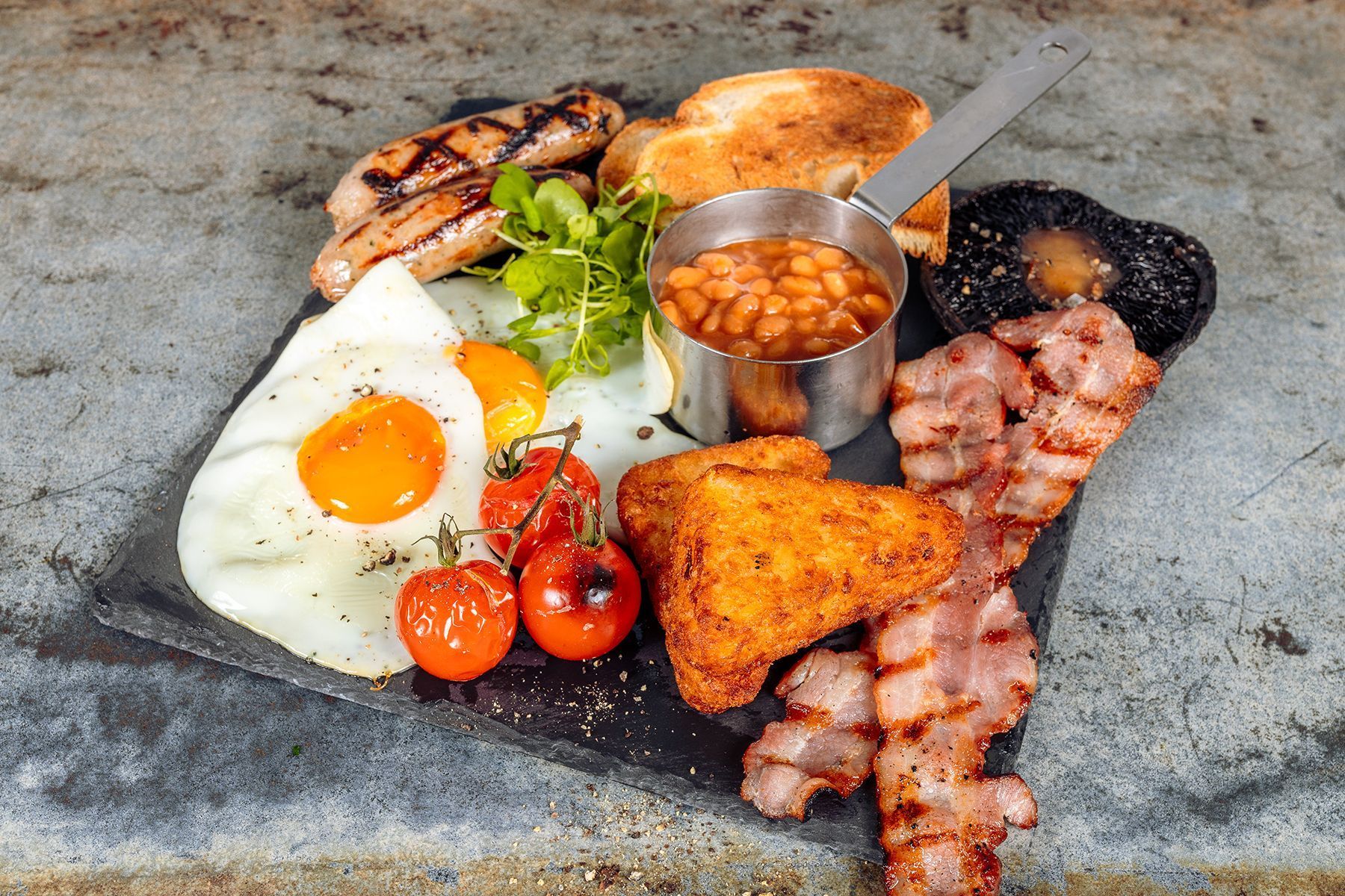 Full English breakfast on a slate: fried egg, bacon, sausage, beans, hash brown, tomatoes, toast.