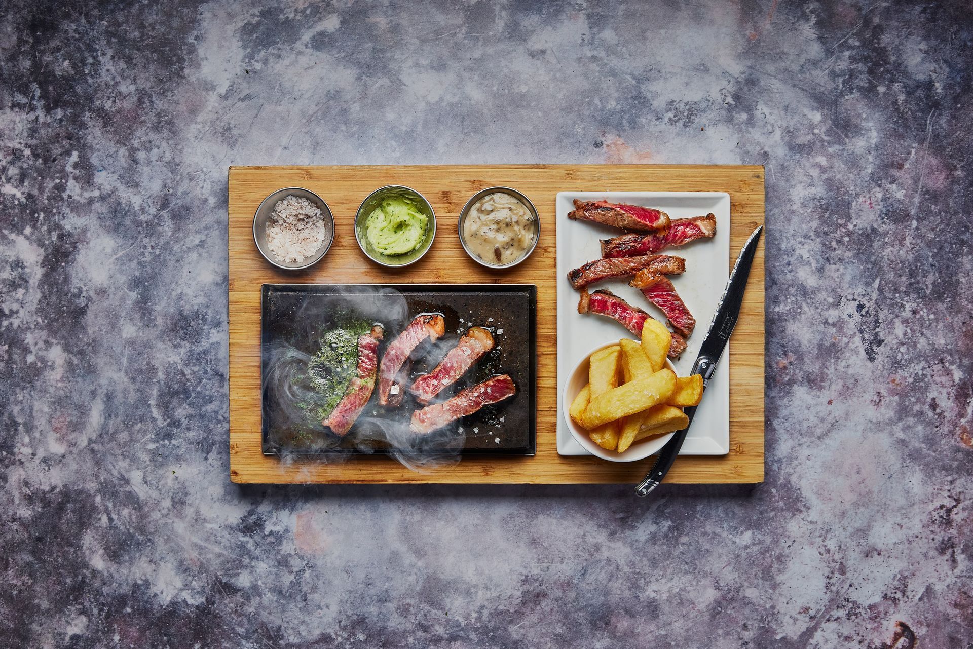 Steak meal on a wooden board with a hot stone, side dishes, and dipping sauces.