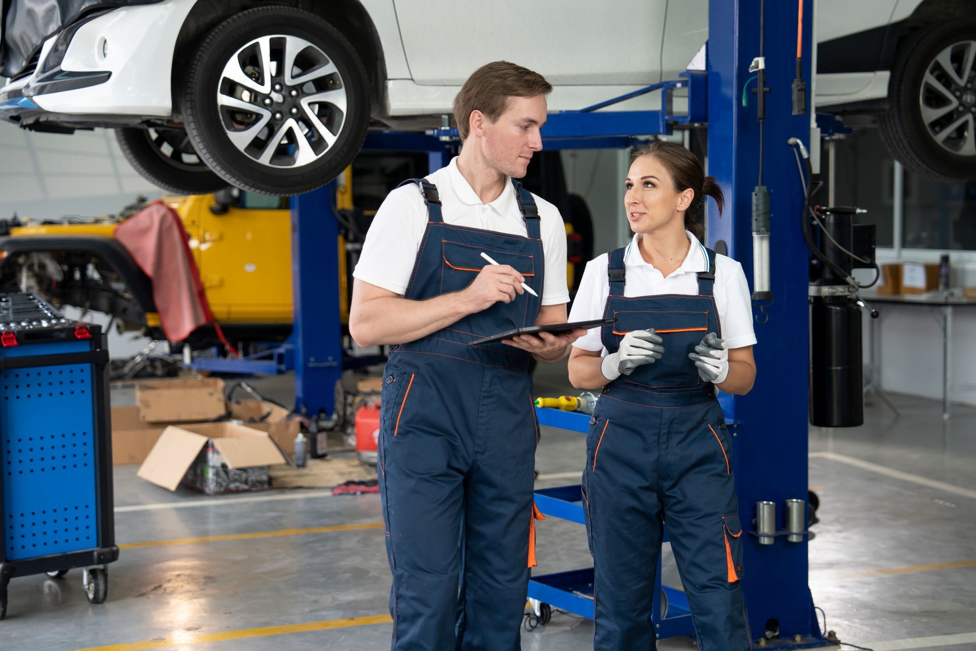 Two mechanics in a garage, discussing paperwork with a car lifted in the background.
