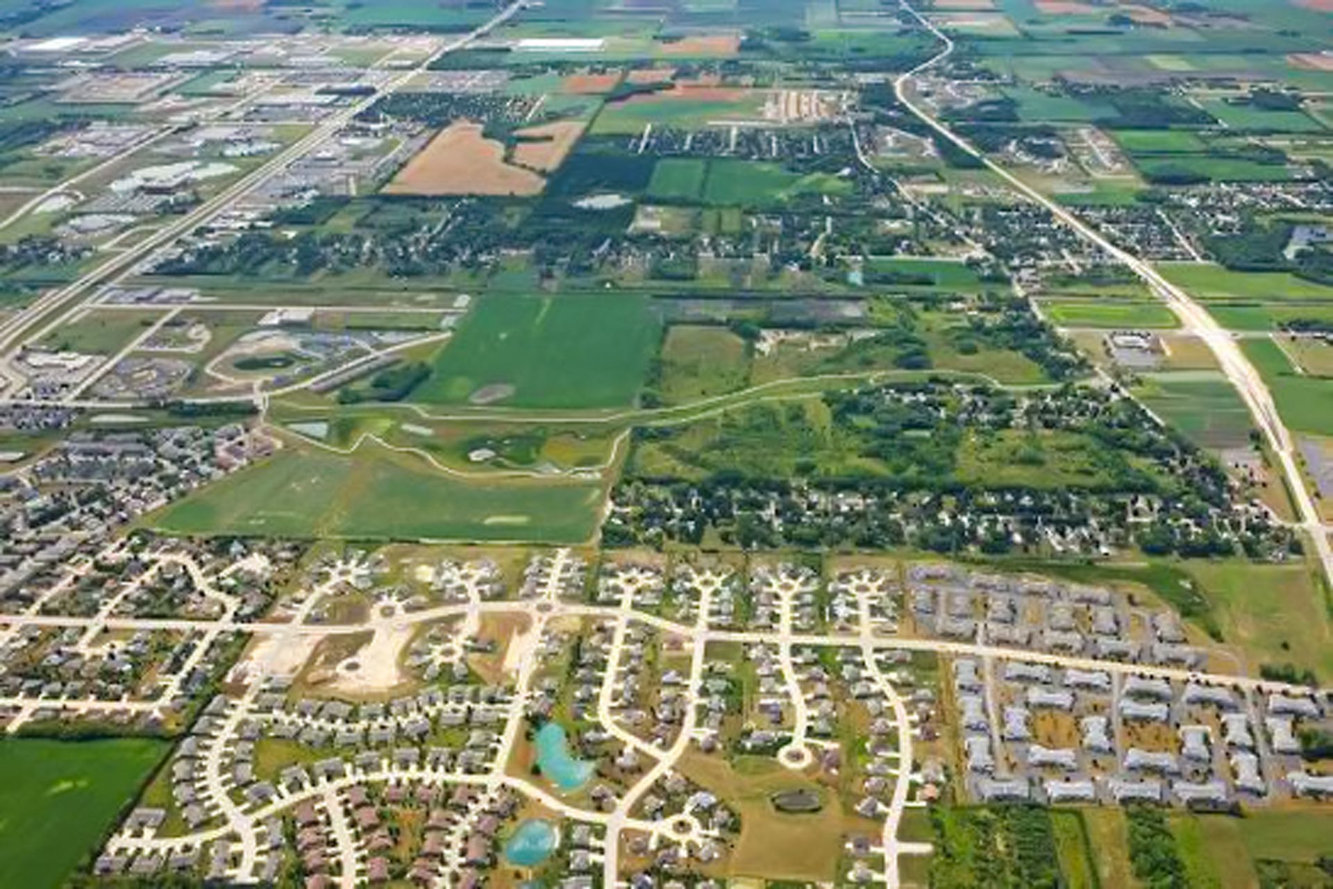 Aerial view of a suburban neighborhood beside farmland, with roads, houses, and green fields