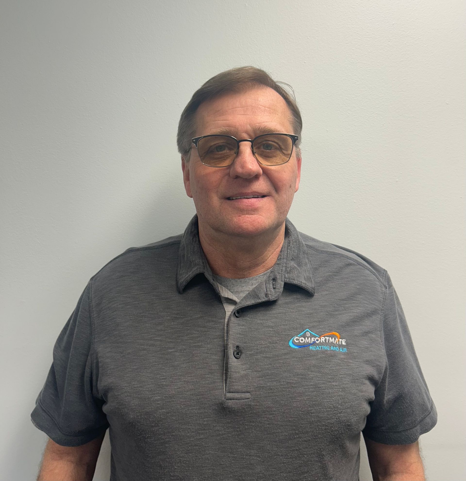 A man wearing glasses and a grey shirt is standing in front of a white wall.