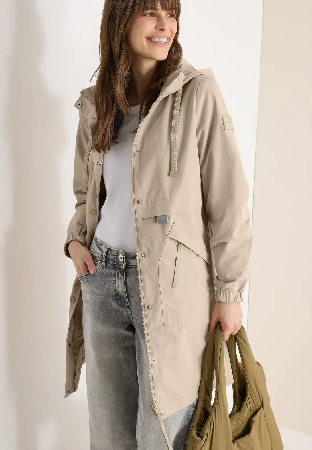 Donna in cappotto marrone chiaro e jeans con in mano una borsa verde, in piedi in un interno.