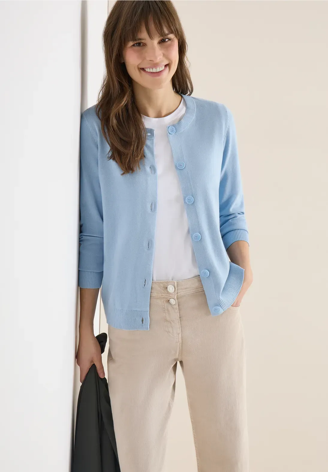 Donna con cardigan azzurro e pantaloni beige, sorridente, appoggiata a un muro bianco.