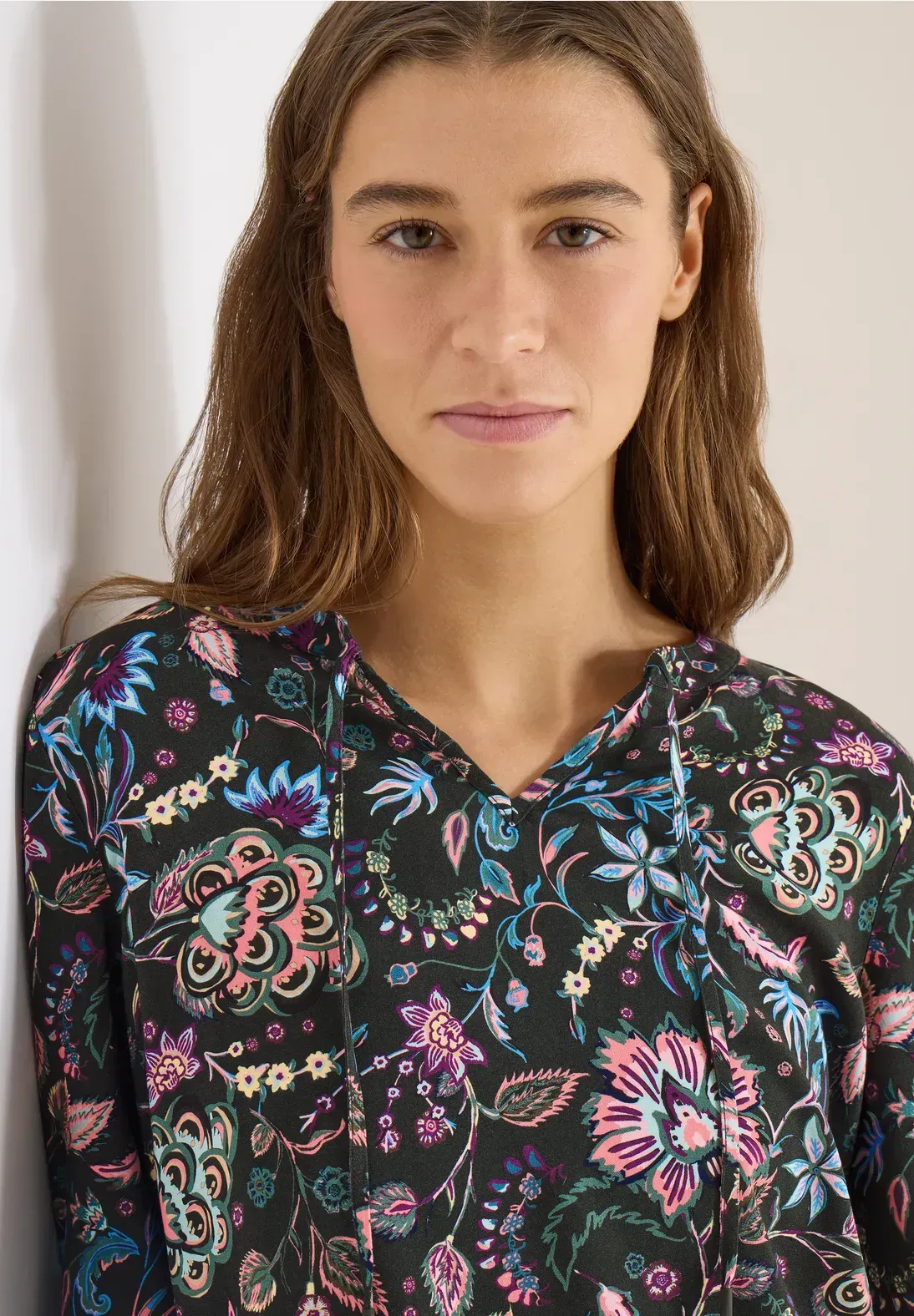 Donna che indossa un top nero con stampa floreale, appoggiata a un muro bianco.