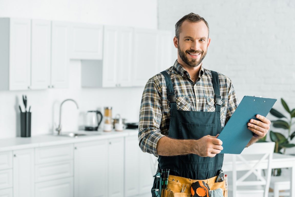 A smiling worker in a plaid shirt and tool belt holds a clipboard in a bright, modern kitchen.