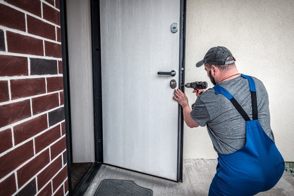 A worker wearing blue overalls uses a power drill to install a lock on a white door in an outdoor doorway.