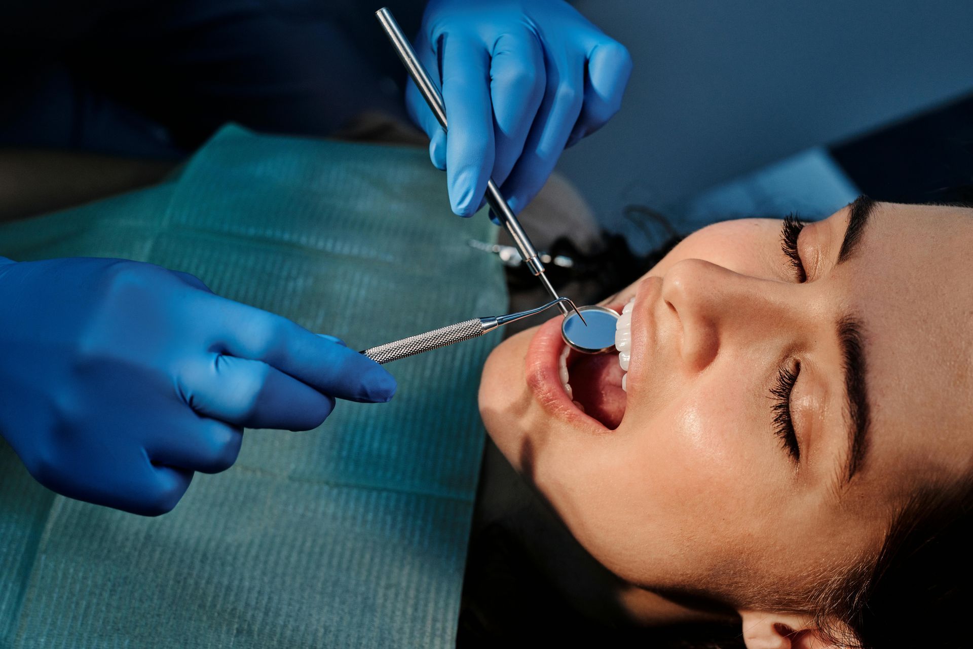 Dentist examining a patient's open mouth with mirror and probe; blue gloves, dental setting.