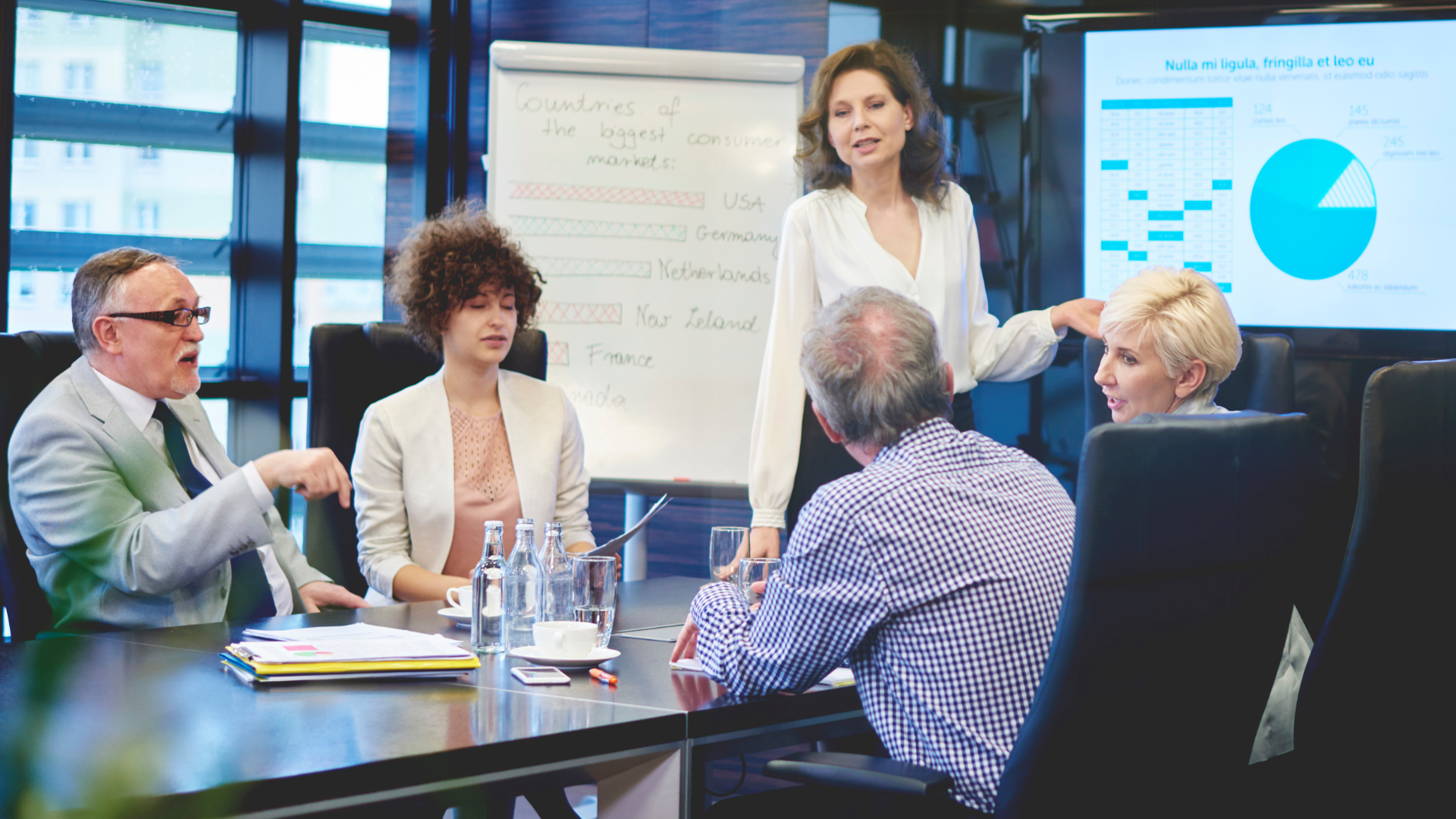 A group of people sit around a boardroom table while a woman stands by a screen and whiteboard during a presentation.