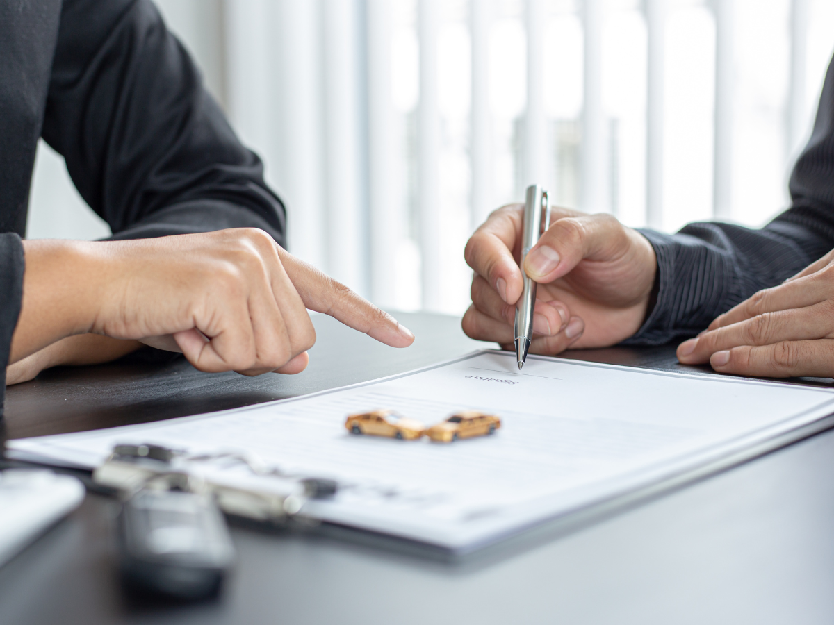 Person signing document with pen, another person pointing at the document; car keys and toy car on desk.