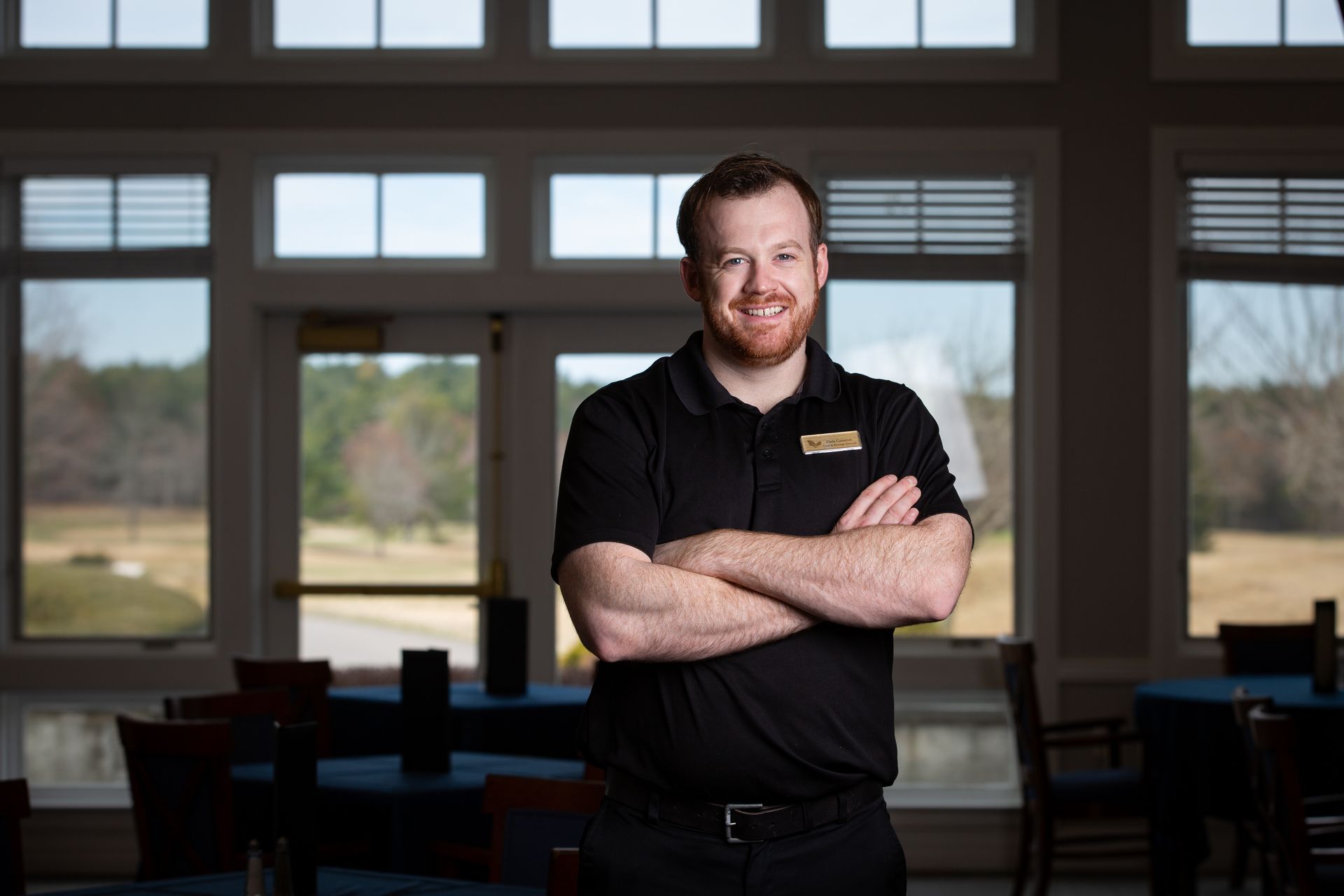 A man with a beard wearing a black under armour polo shirt is standing in front of a window.