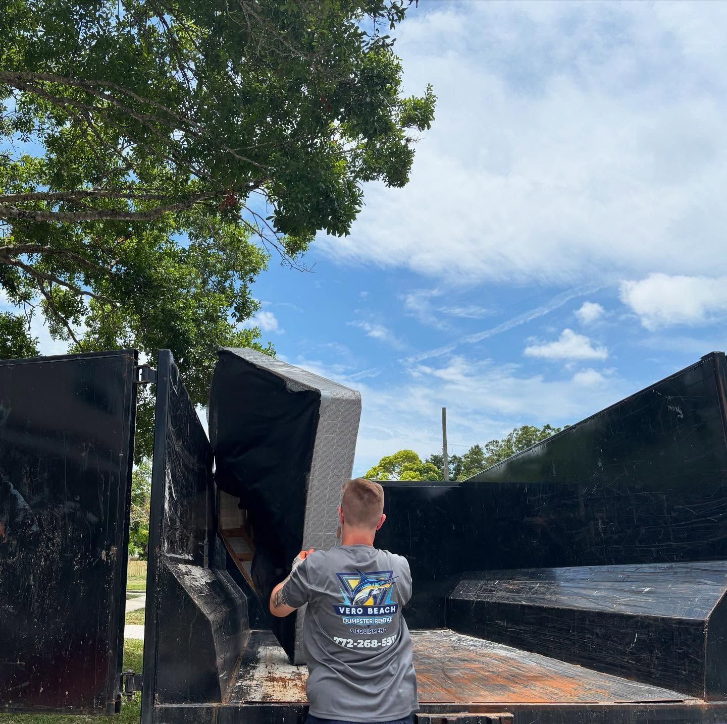 Man in grey shirt loading a piece of furniture into a dumpster under a bright blue sky.