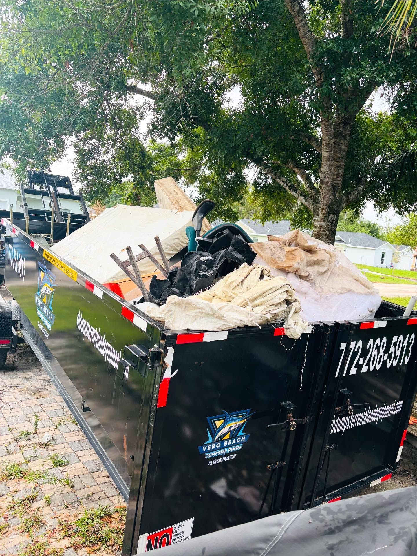 Man thumbs-up by a black dumpster filled with debris. Outdoor setting, clear sky. Phone number visible.