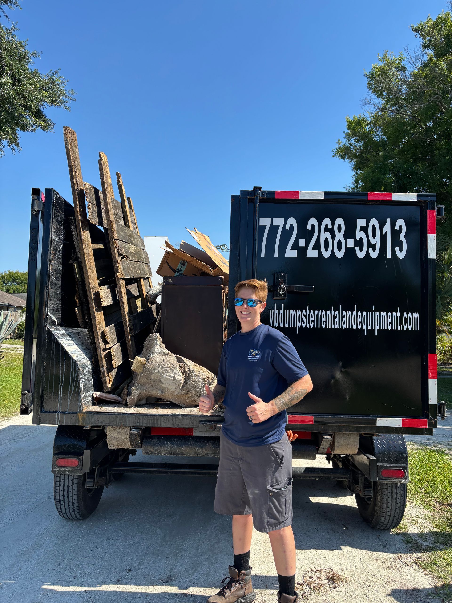 Man thumbs-up by a black dumpster filled with debris. Outdoor setting, clear sky. Phone number visible.