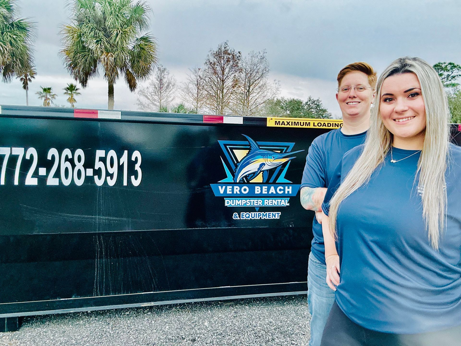 Two people stand in front of a Vero Beach dumpster. They are smiling and wearing blue shirts.