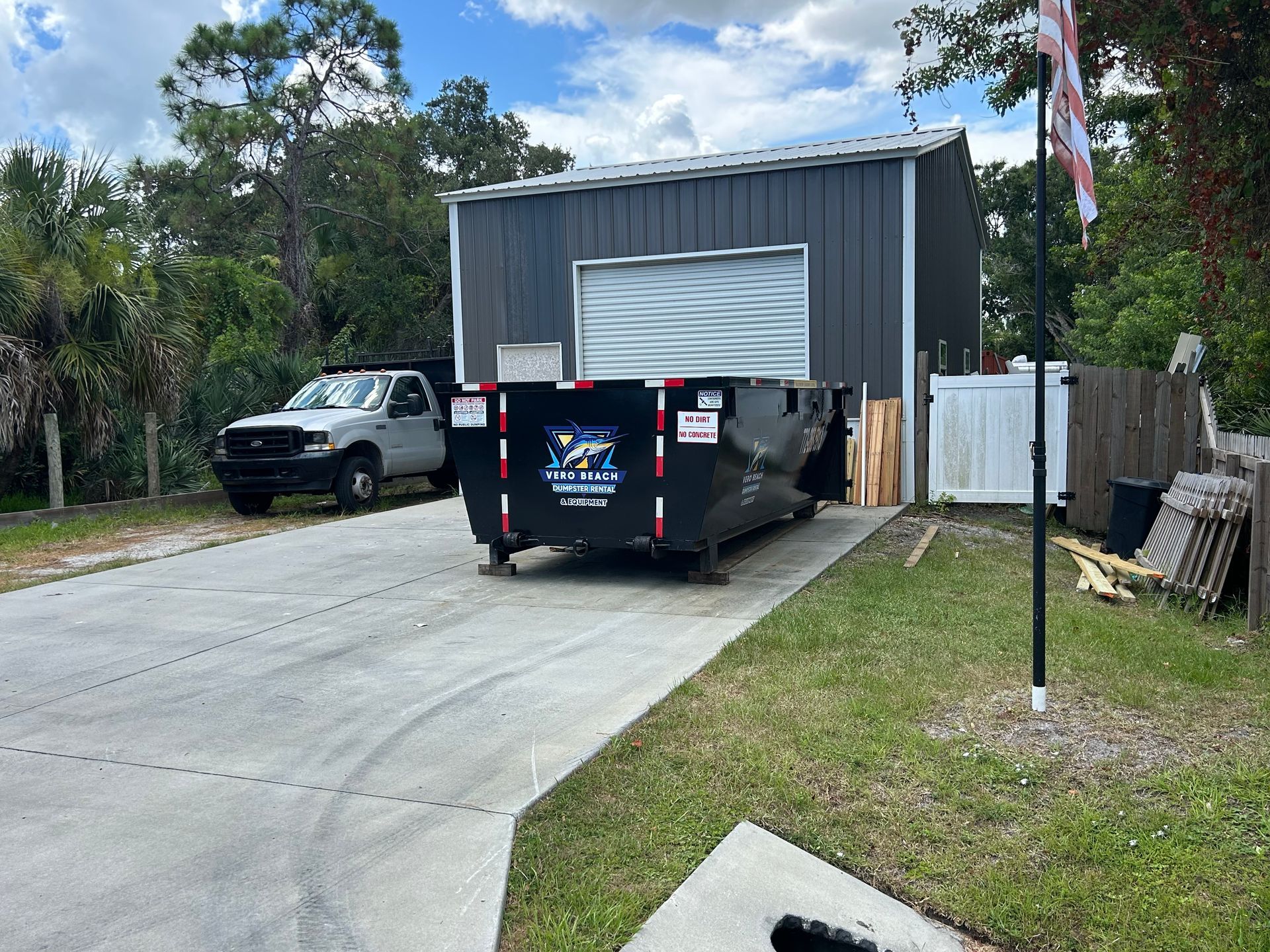 A black dumpster in a driveway with a pickup truck and metal building.