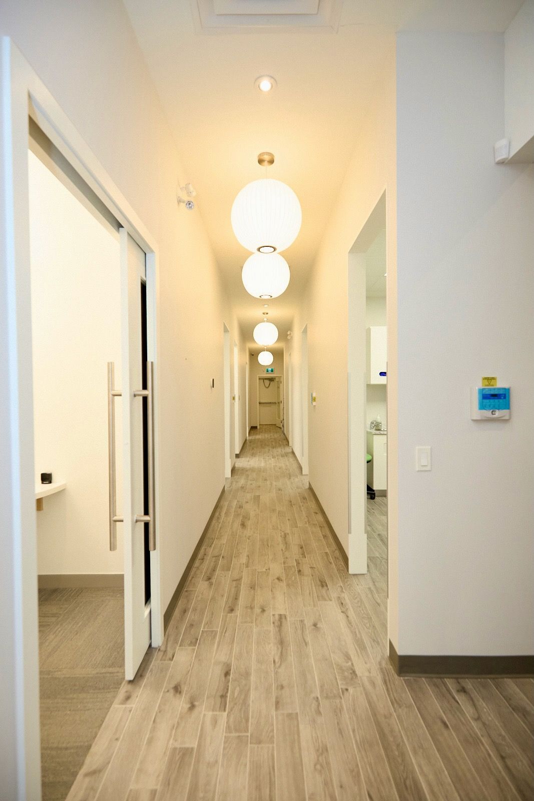 Long hallway with light wood flooring, white walls, and globe pendant lights. Open doors line the sides.