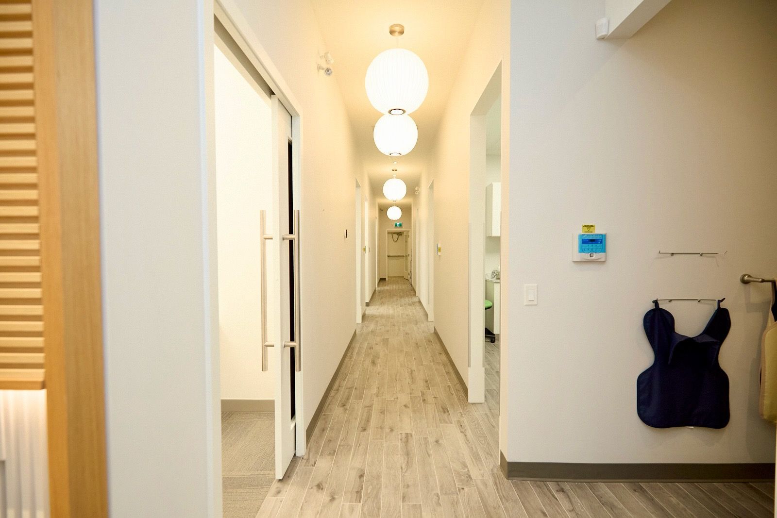 Long, bright hallway with round lights, wooden floor, open doorways, and hanging medical apron.