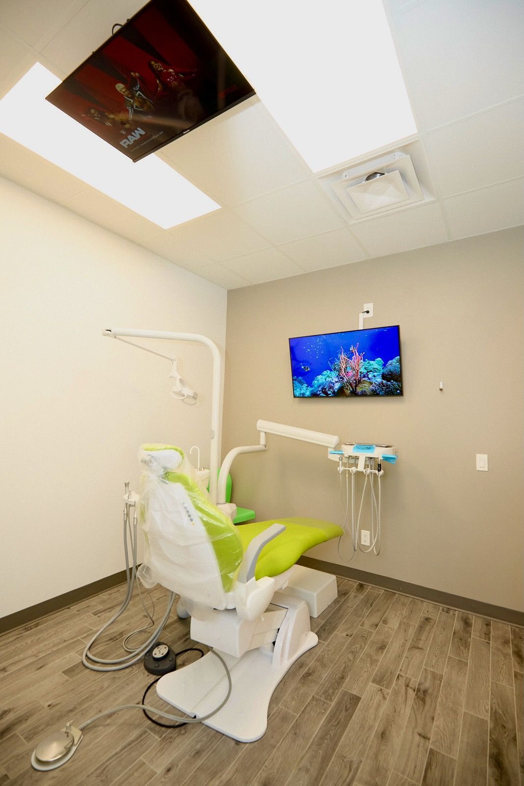 Dentist's office with green dental chair, tools, TV showing an aquarium scene, recessed lights, and gray wood-look floor.