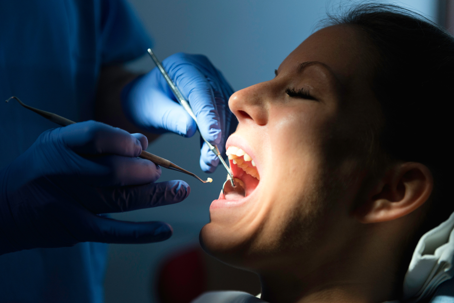 Dentist examining a patient's open mouth with dental tools. Dark blue setting.