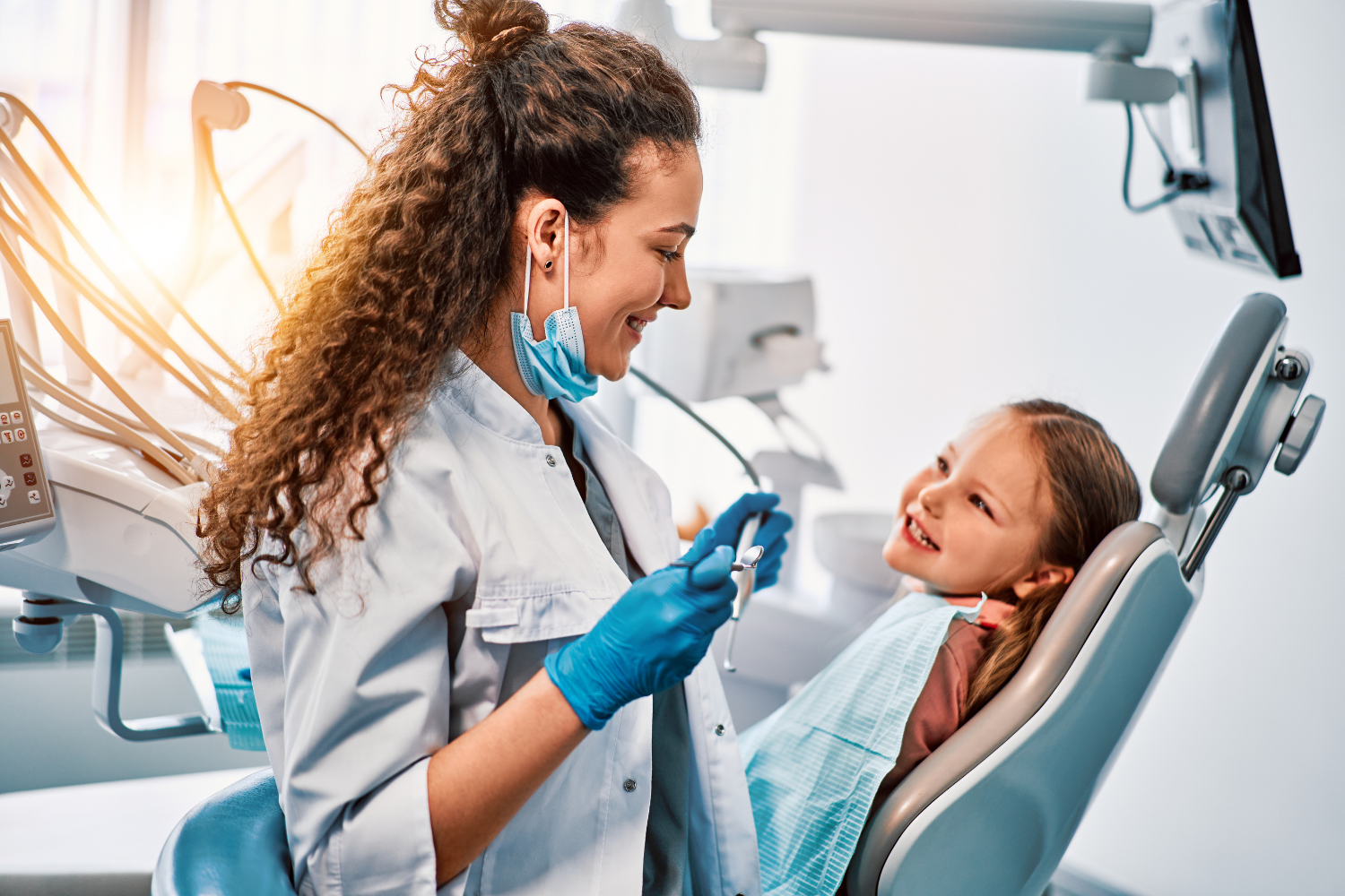 Dentist smiling at child in dental chair, holding tools. Bright office setting.