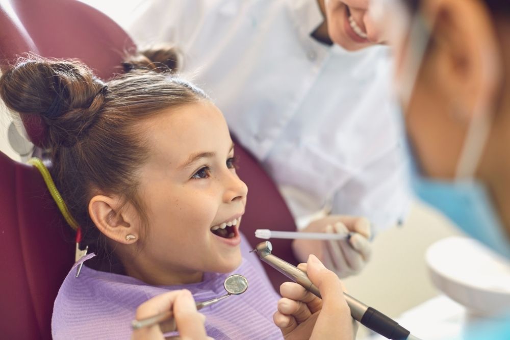 Child smiles wide in a dentist's chair while a dentist works on her teeth.