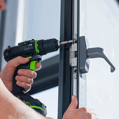 A Locksmith Uses A Drill To Repair A Commercial Lock On A Glass Door.