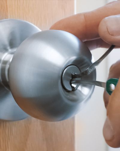 A Technician Uses Two Lock Picks To Open A Doorknob On A Wooden Door.
