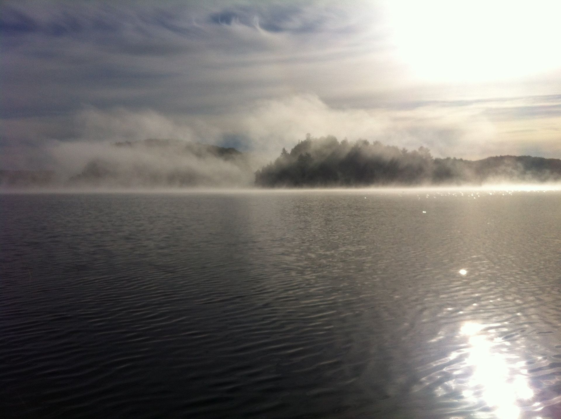 The sun is shining through the clouds over a foggy lake.