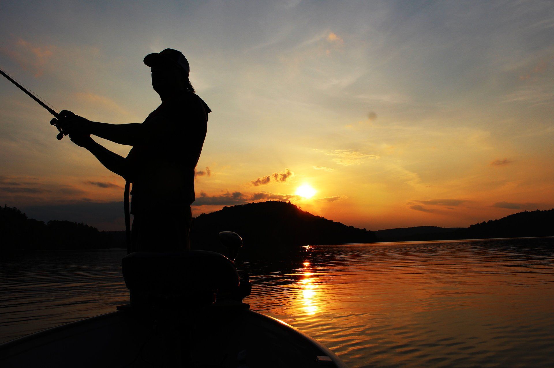 A man is fishing from a boat at sunset.