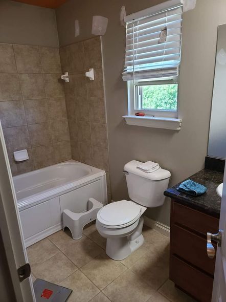 Bathroom with tub, toilet, and vanity. Tan tile, white fixtures, gray walls. A small step stool sits by the toilet.