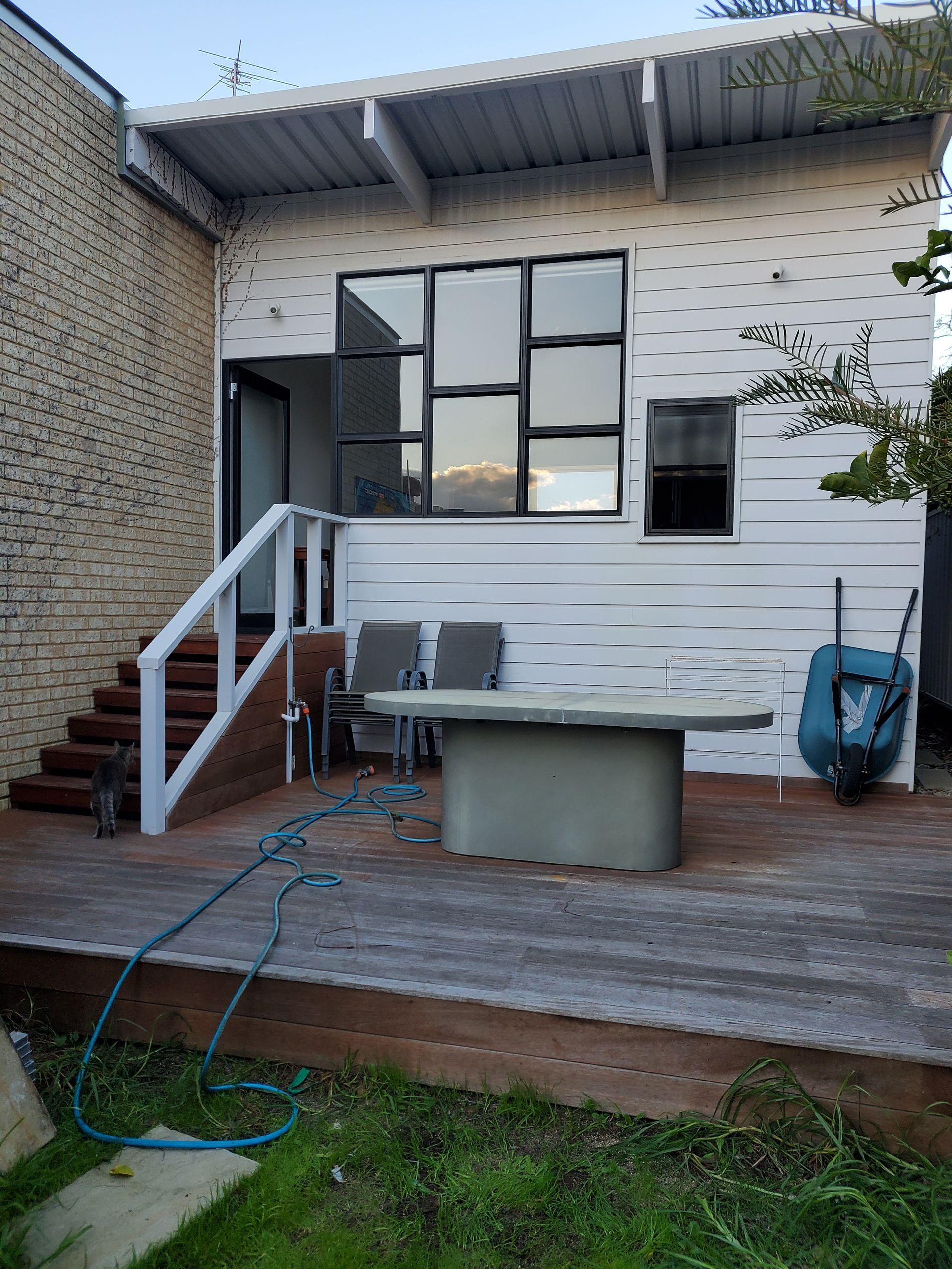 Wooden deck with a white-paneled building, large window, stairs, and a tub. A blue hose lies on the deck.