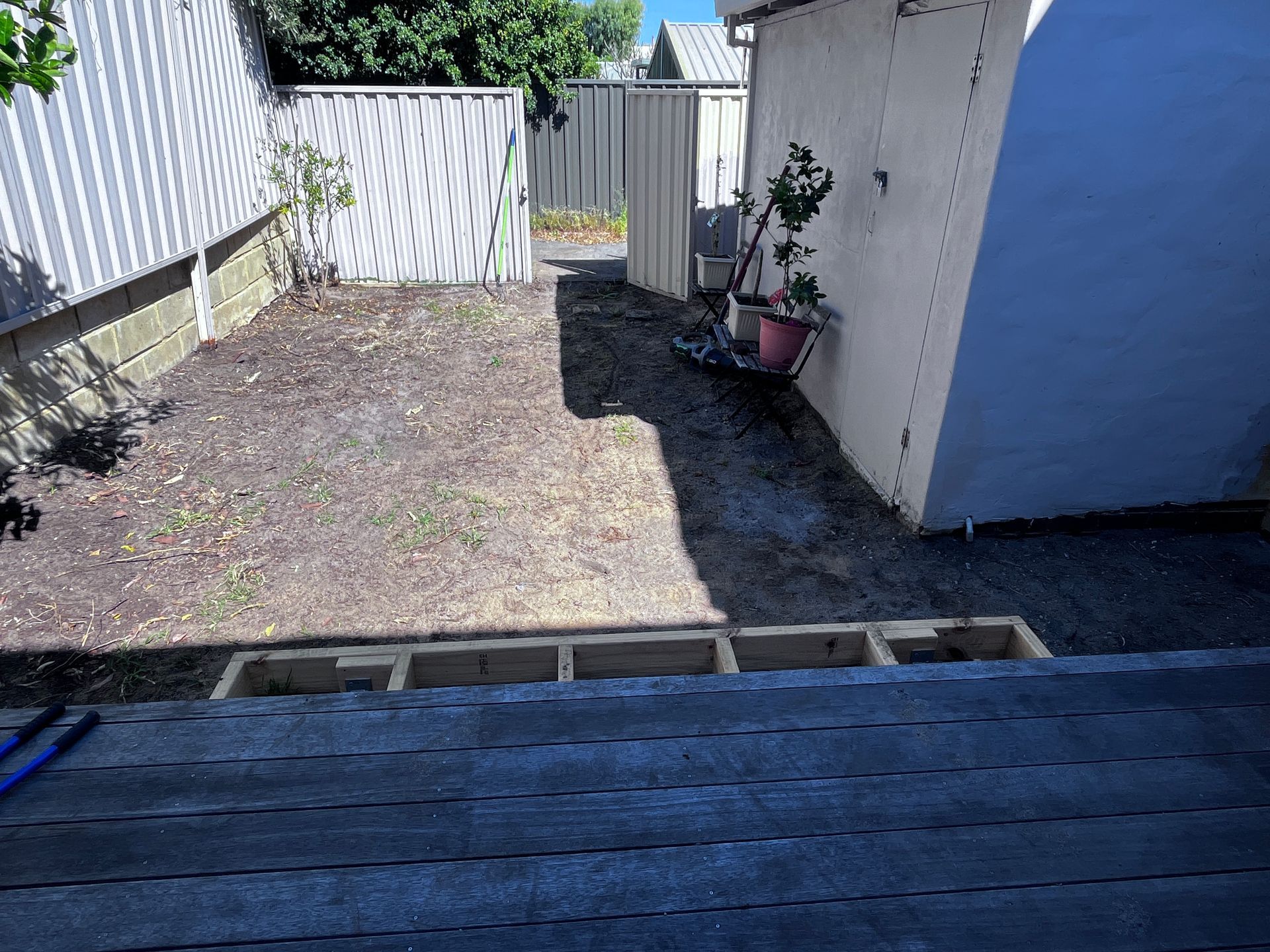 Backyard with a gravel path, dirt, and a wooden deck frame. Fences and a building are in the background.