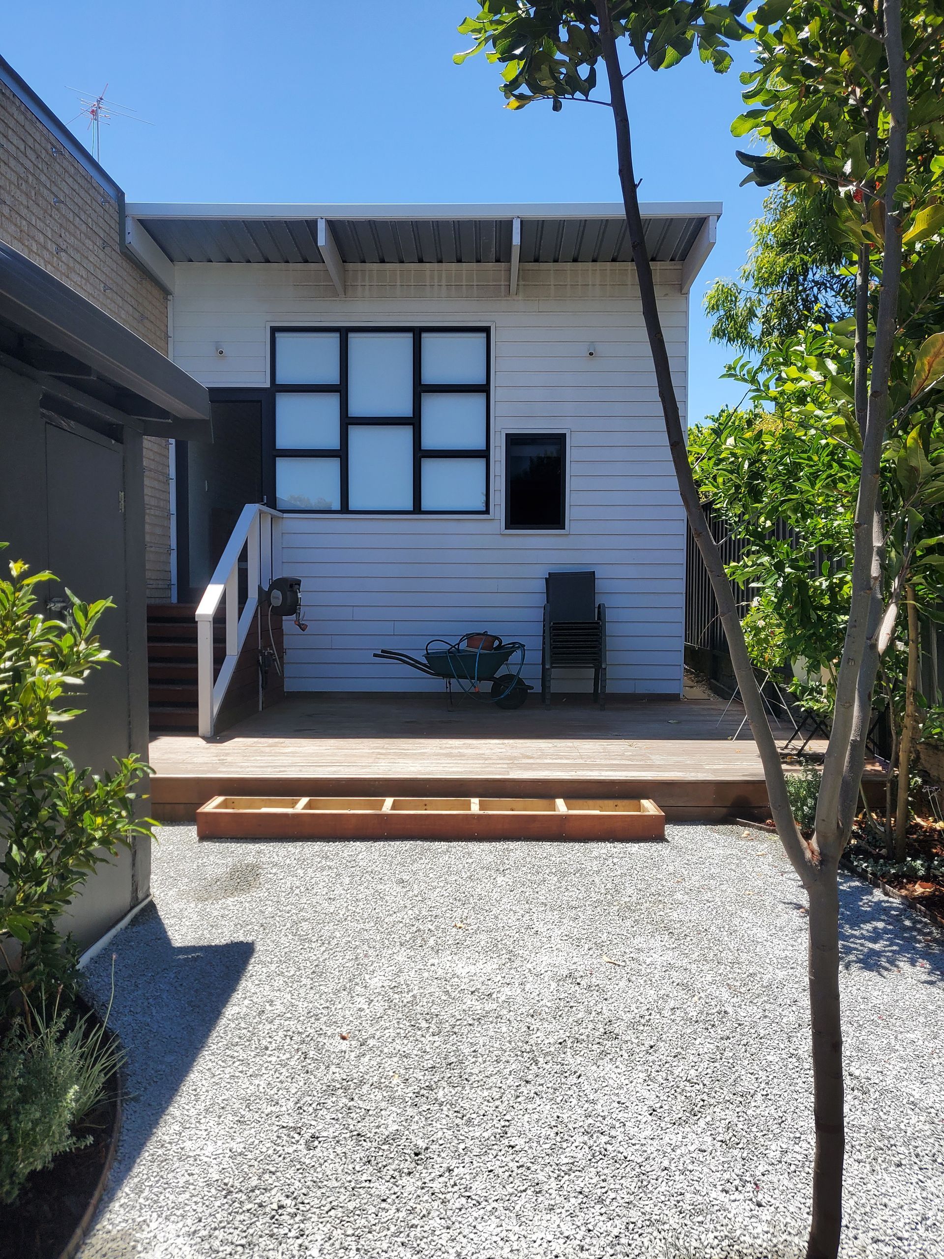 White clapboard building with windows, a deck, gravel yard, and trees.