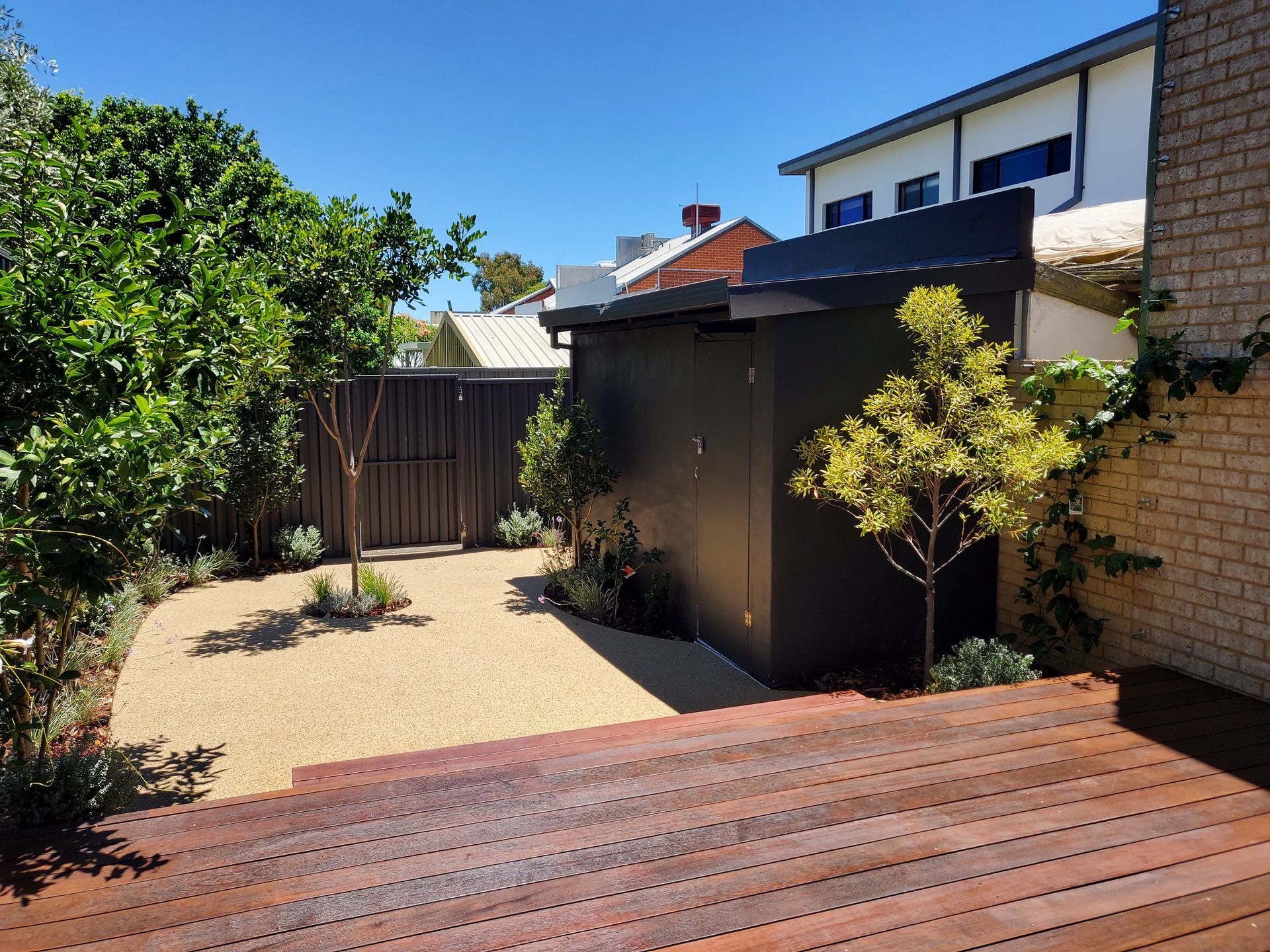 Backyard with wooden deck, gravel path, black shed, and lush greenery under a clear blue sky.