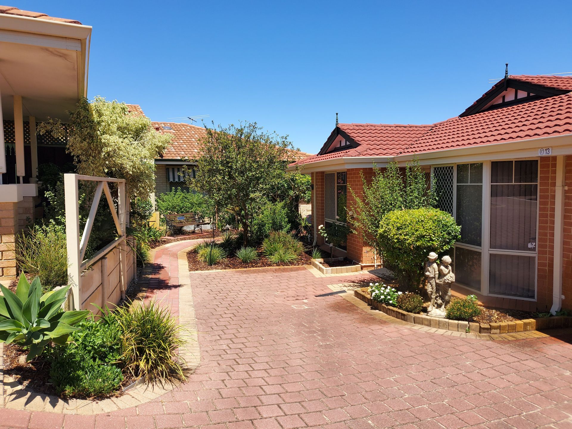 Brick pathway between red-roofed brick buildings with lush greenery under a clear blue sky.