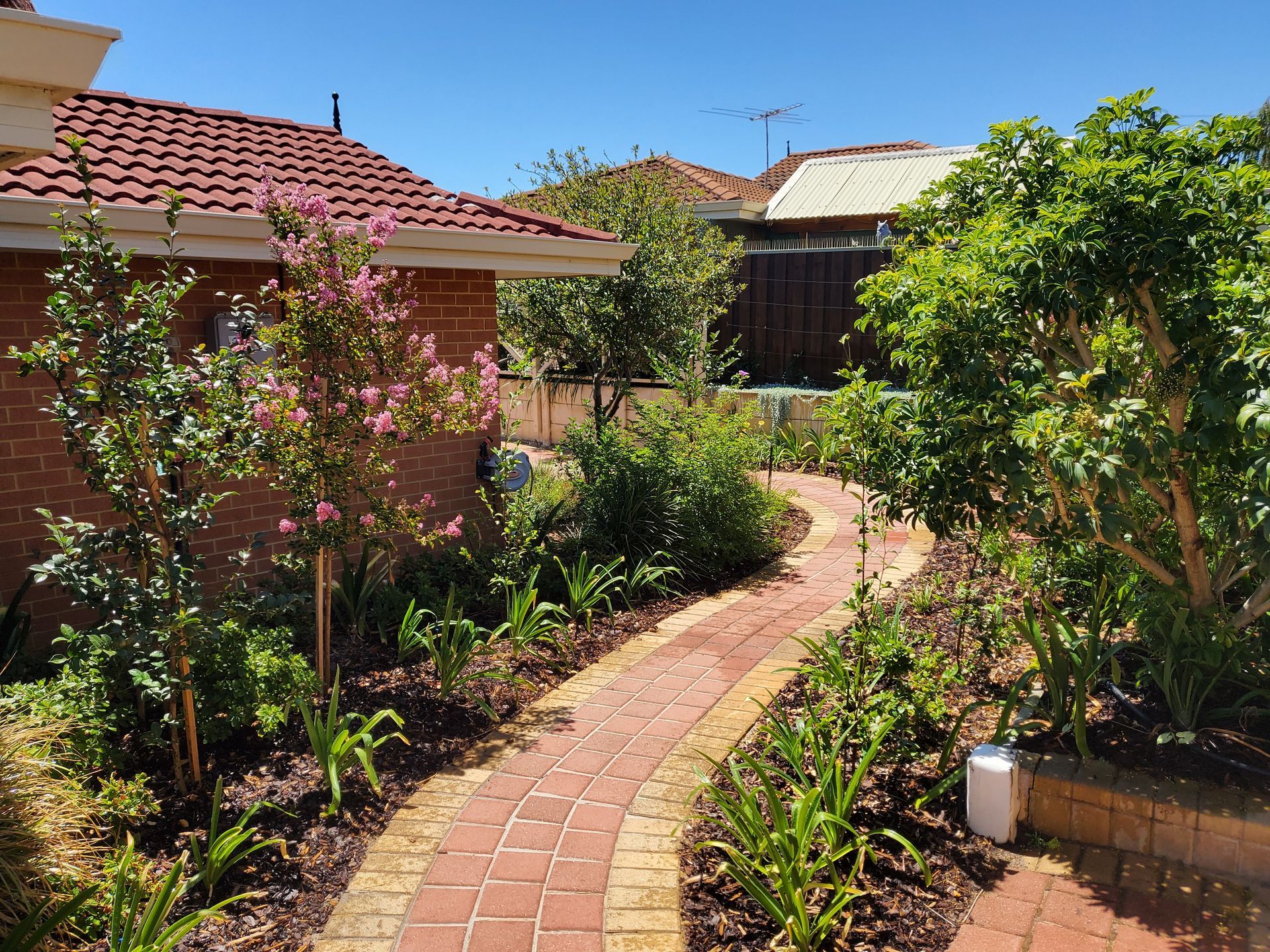 Brick pathway curves through a lush garden alongside a red brick house with a tiled roof.