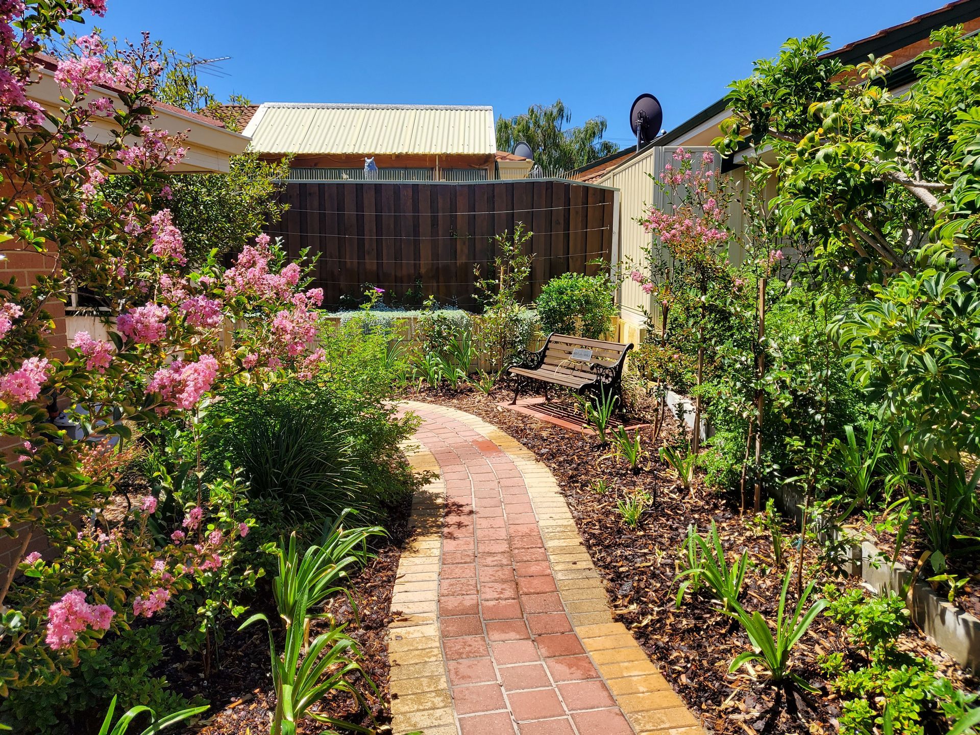 Brick path winds through a garden with blooming pink flowers, lush green foliage, and a wooden fence.